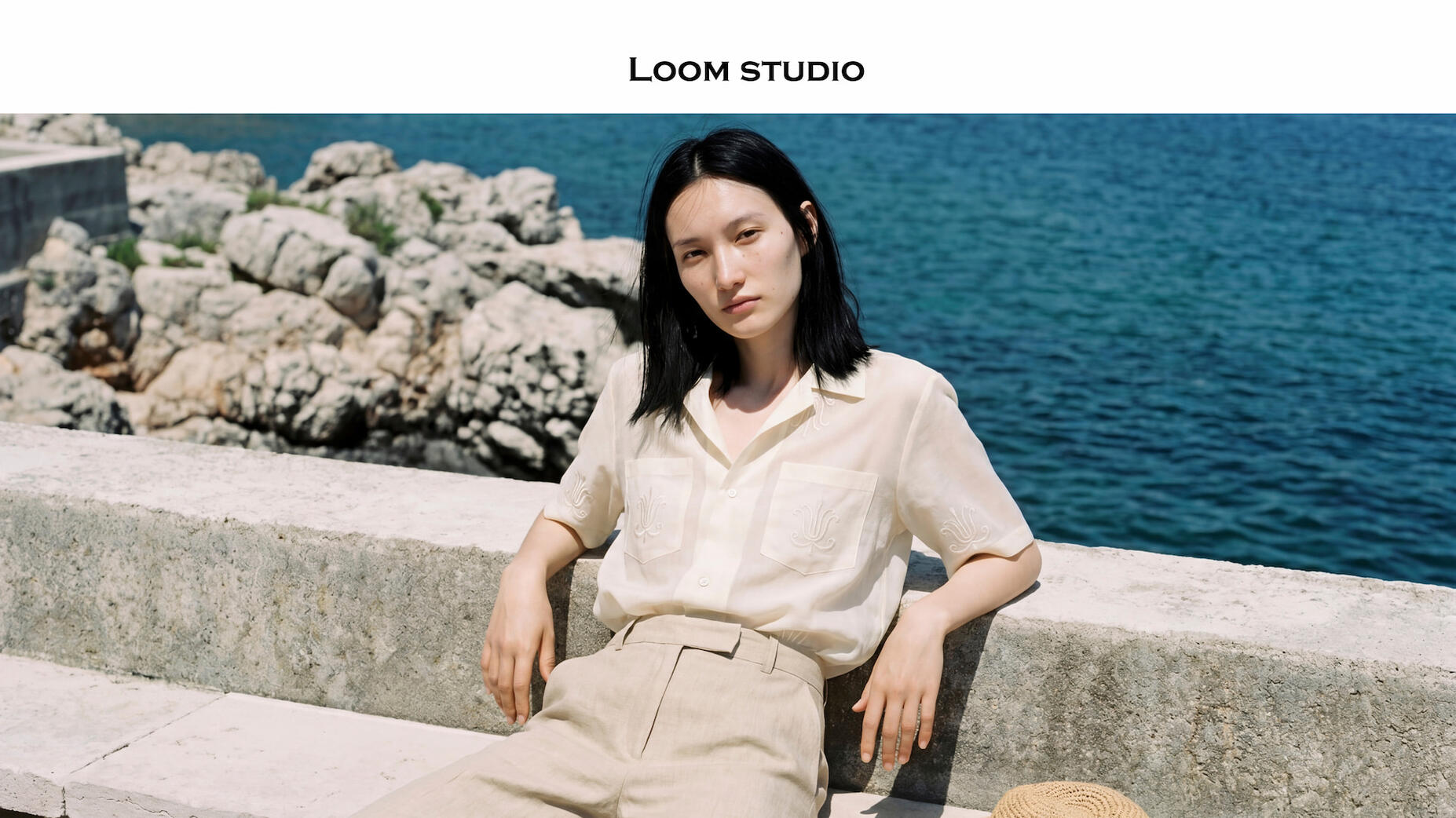 Riviera Reverie: Translucent Silk and Embroidered Details AI-generated fashion photography by Loom Studio featuring a model in a sheer cream embroidered short-sleeve shirt and beige linen trousers. Coastal Mediterranean background with sea and rocks, showcasing high-fidelity embroidery and translucent fabric rend
